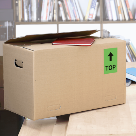 Kartonbox auf einem Tisch, mit einem roten Heft und einem grünen "TOP"-Schild auf der Seite. Im Hintergrund sind Bücherregale sichtbar.