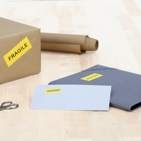 Ein Karton mit dem Aufkleber "FRAGILE", daneben ein graues Kuvert mit dem Aufkleber "URGENT!" und eine Schere auf einem Tisch.